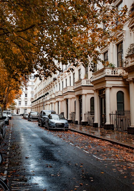 A street view of elegant white residential buildings with classic architectural details, including arched windows, decorative moldings, and black wrought iron balconies. A leafless tree stands promine